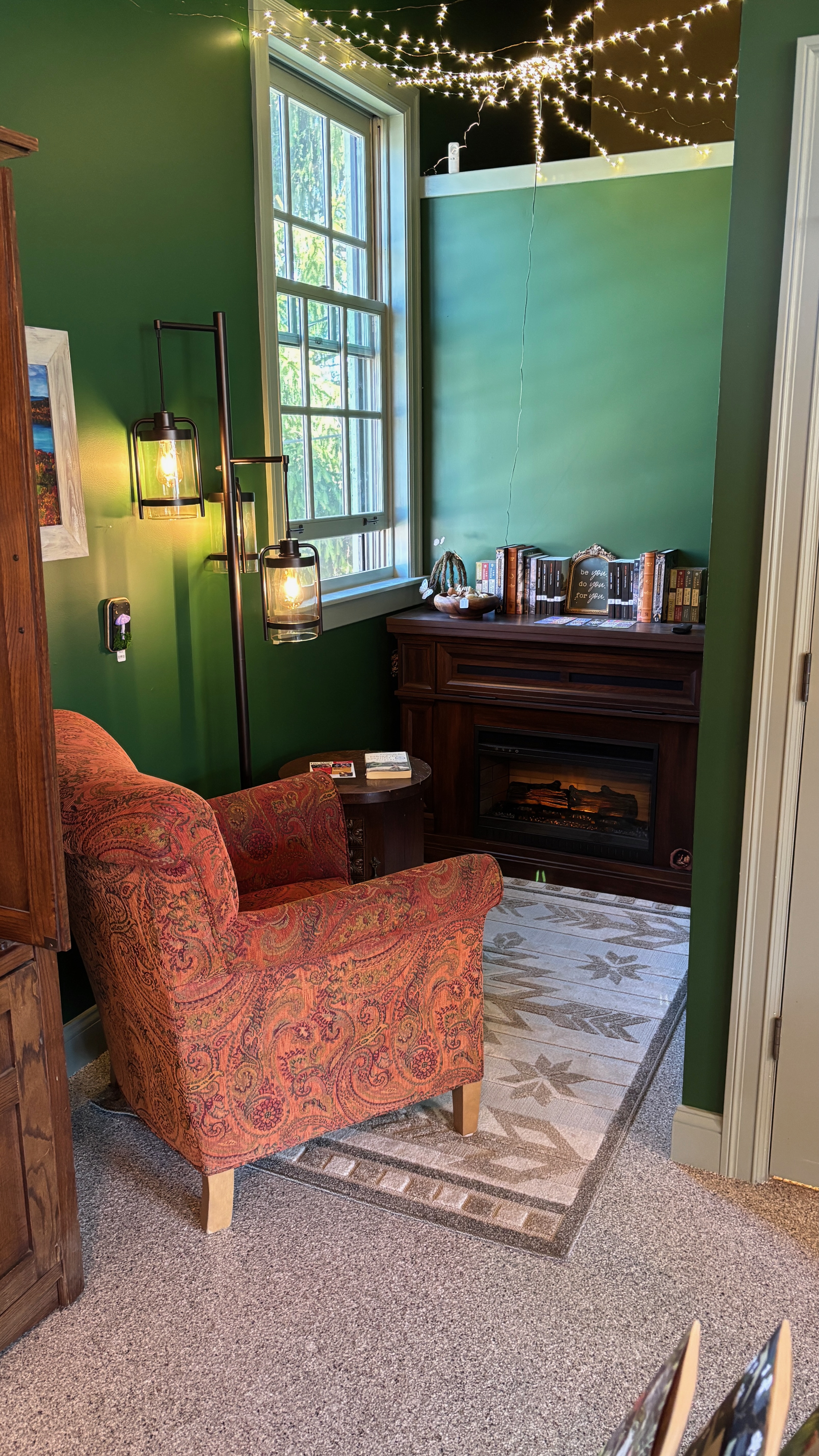 A nook in a book shop, about 1.5 meters square with a fireplace in the back, a large window on one wall, and a big comfy chair facing into the nook. There are tiny white lights on the ceiling.