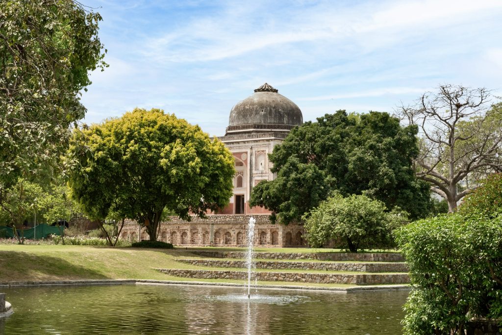 A serene garden scene featuring a peaceful water feature with a fountain in the foreground, surrounded by lush greenery. In the background, a historical building with a large dome is partially obscured by trees, set against a blue sky with wispy clouds.
