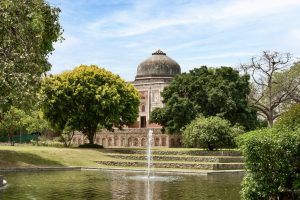 A serene garden scene featuring a peaceful water feature with a fountain in the foreground, surrounded by lush greenery. In the background, a historical building with a large dome is partially obscured by trees, set against a blue sky with wispy clouds.
