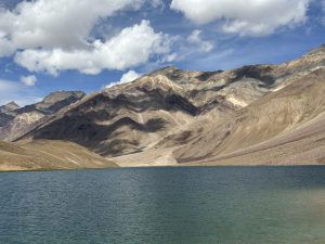A serene landscape featuring a clear blue lake in the foreground, surrounded by rugged, brown and gray mountains under a partly cloudy sky