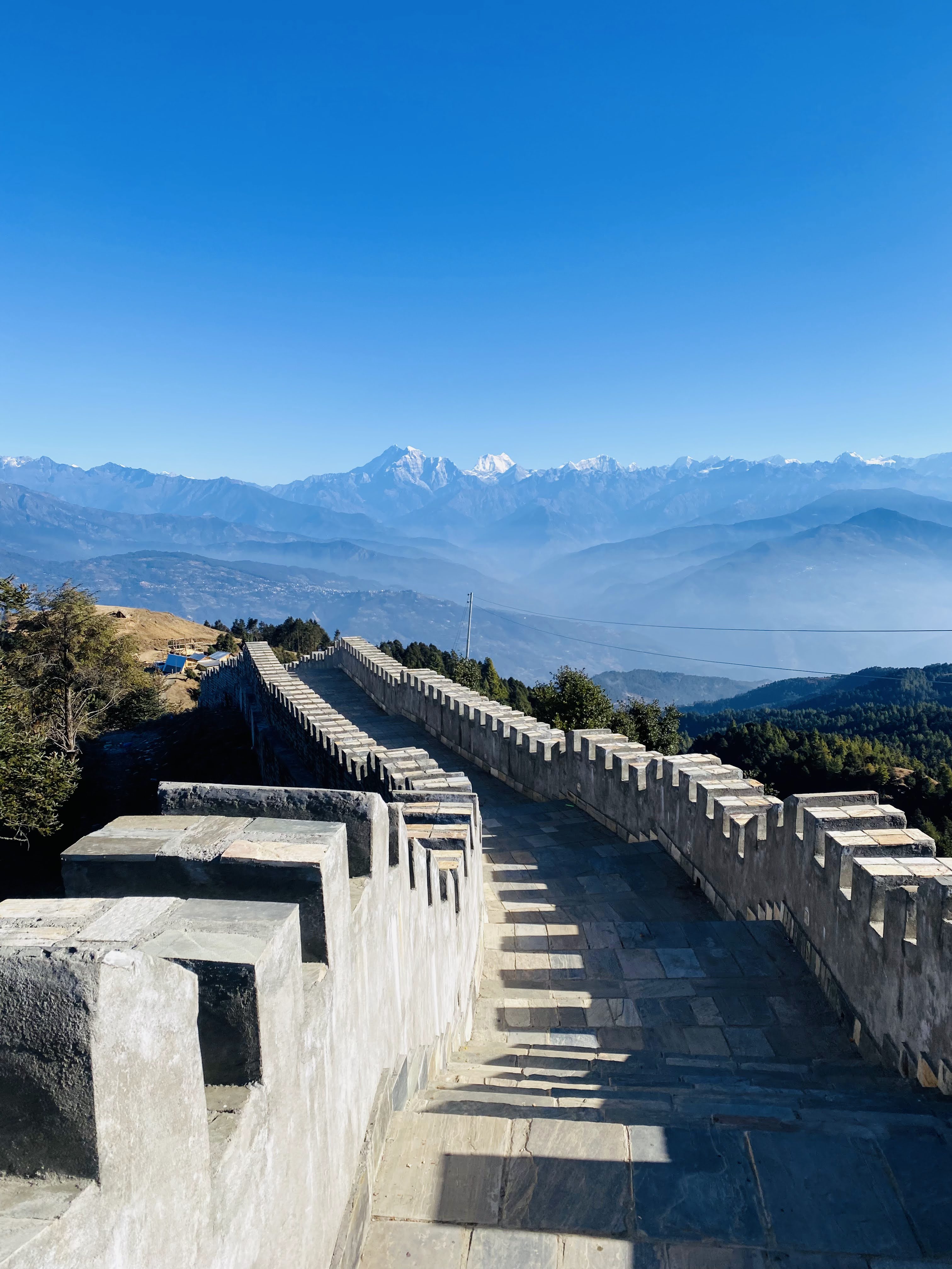 A stone pathway winds along a fortified wall with crenellations, leading toward distant snow-capped mountains under a clear blue sky.