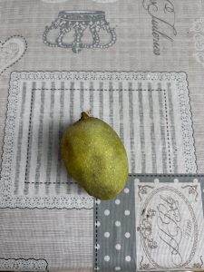 A single, unripe mango with a greenish-yellow skin sits on a textured tablecloth featuring various patterns, including stripes and polka dots in soft gray tones.