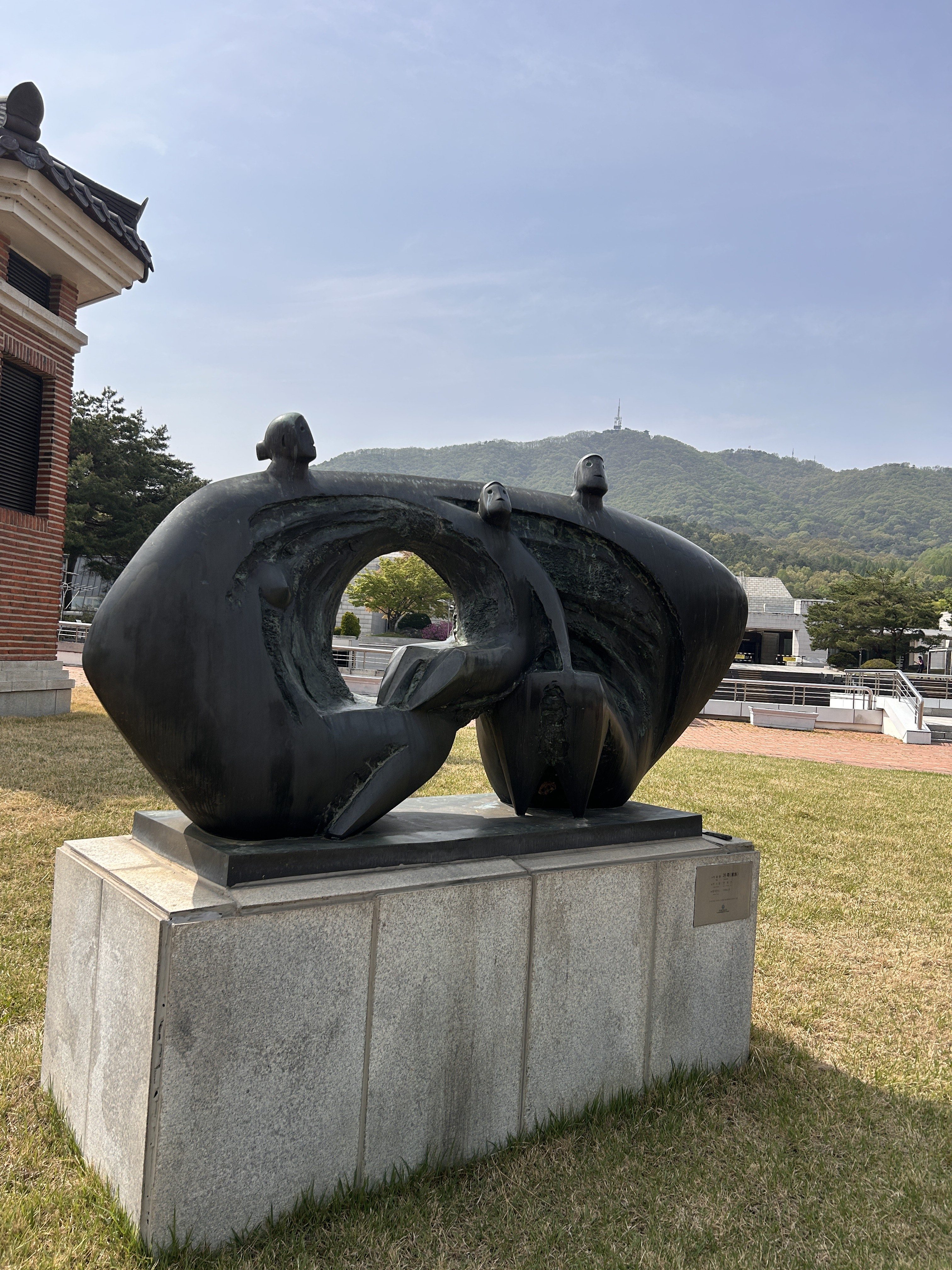 A bronze sculpture featuring three abstract figures. One figure is seated and appears to be interacting with the other two figures, who are positioned above and to the sides. The sculpture is set on a stone base and surrounded by grass, with a backdrop of trees and a distant hill. The sky is clear, and the scene appears peaceful.