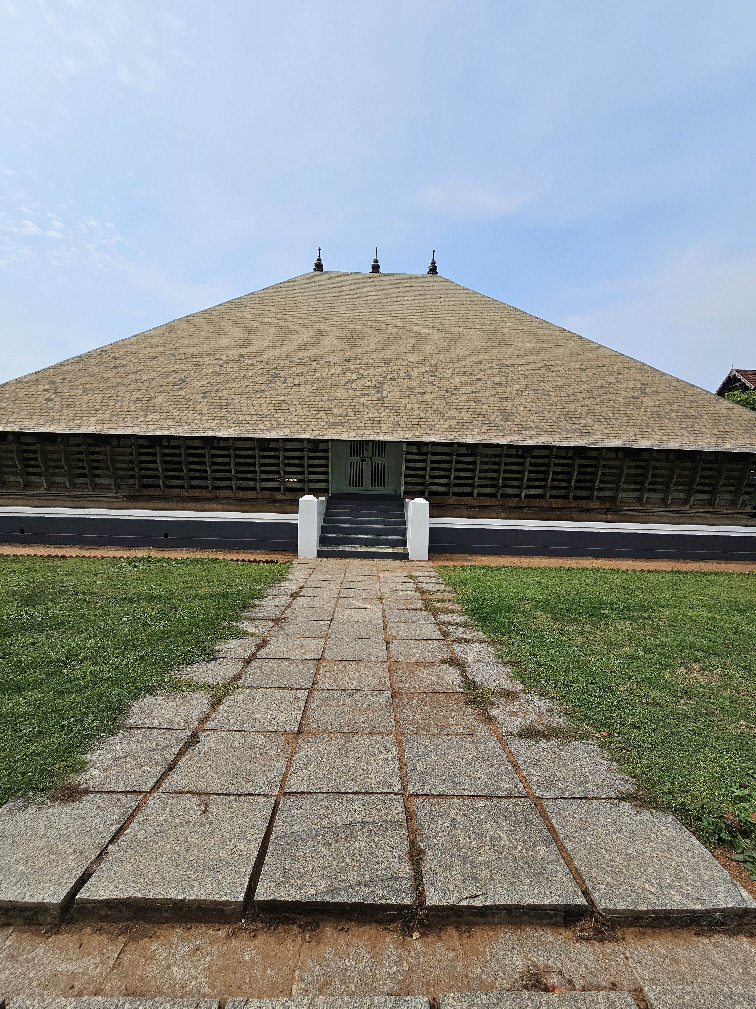 The Koothambalam (temple theatre or play house) of Vadakkunnathan Temple in Thrissur, Kerala, India, features its distinctive sloping roof and traditional Kerala architectural style. A stone-paved pathway leads to the temple entrance, with green grass visible on either side.