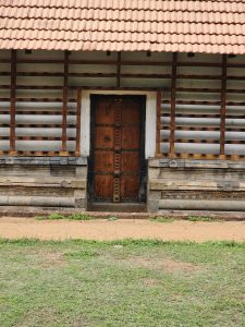 An entrance of the Vadakkunnathan Temple in Thrissur, Kerala. The temple's wooden door is centered, flanked by a white frame and set against a backdrop of stone and wood architecture, with a tiled roof overhead and a mix of grass and sandy ground in the foreground.