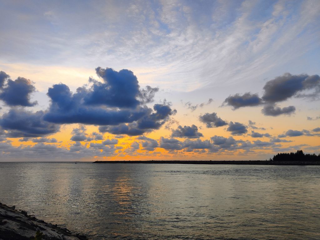 A serene view of a body of water at sunset, with vibrant hues of orange and yellow blending into the blue sky. Fluffy clouds are scattered across the sky, reflecting the warm colors of the sunset on the water’s surface. In the distance, a jetty extends into the water, and a silhouette of trees is visible along the horizon.
