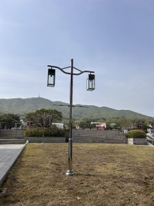 A shiny, modern lamppost with two lanterns stands on a grassy area. In the background, a low green mountain is visible under a clear blue sky.