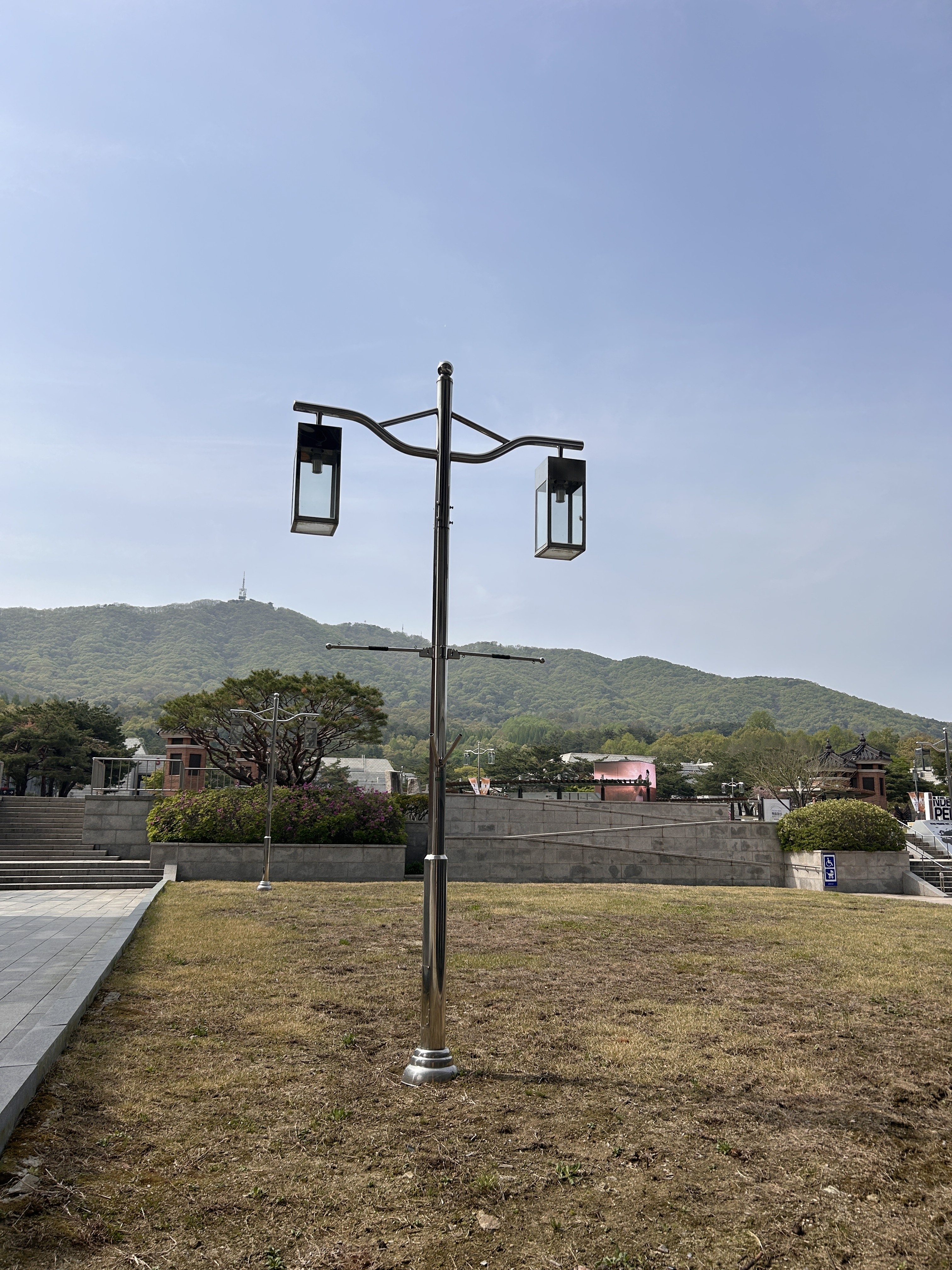 A shiny, modern lamppost with two lanterns stands on a grassy area. In the background, a low green mountain is visible under a clear blue sky.