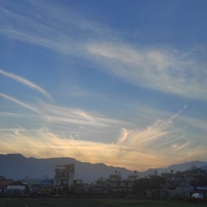 High wispy clouds in a crepuscular sky, low mountains on the horizon, buildings in the foreground.