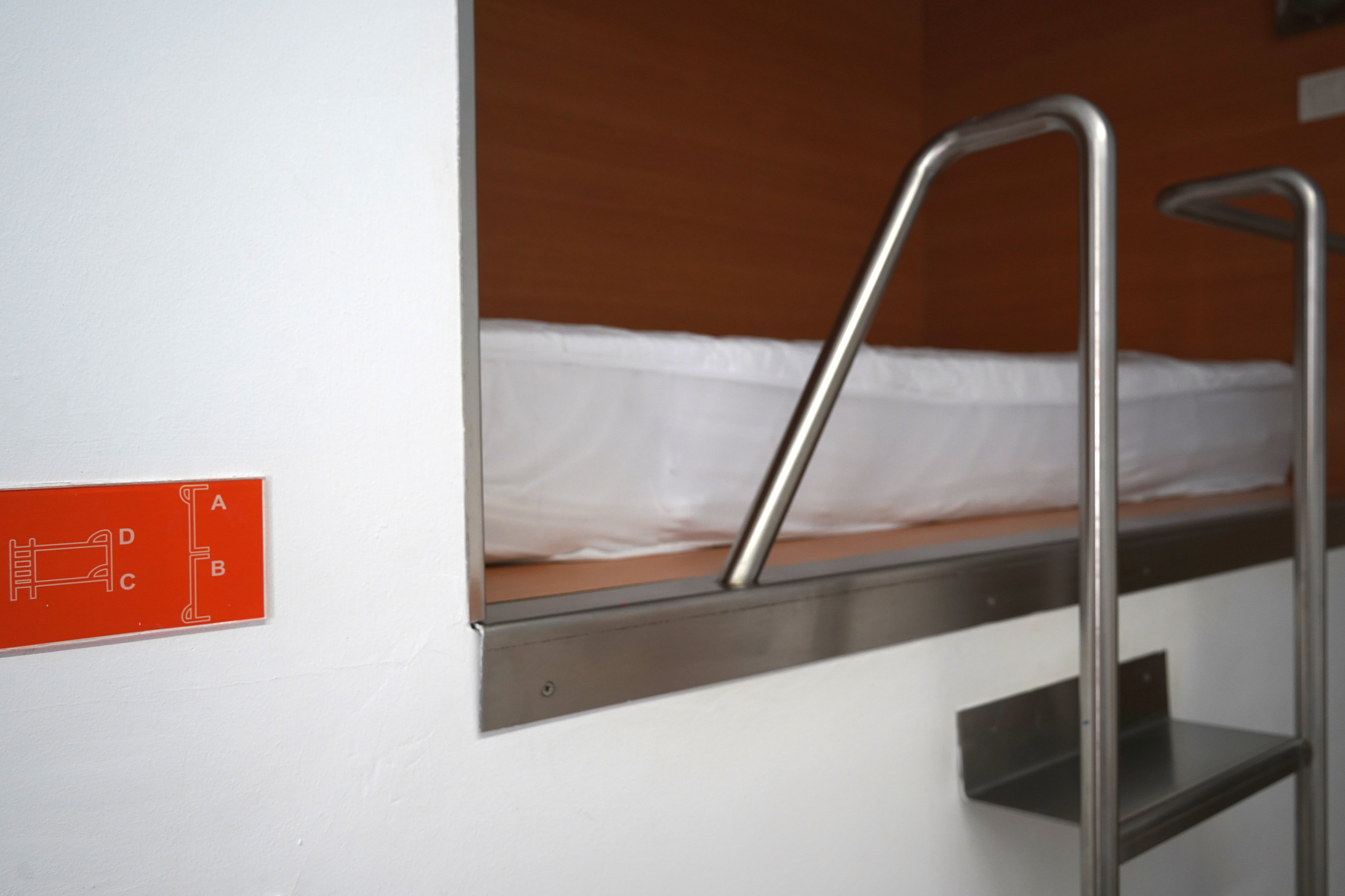 An interior view of a compact sleeping space featuring a raised bed with a white mattress and a metal safety railing. To the left, there is an orange signage panel with white illustrations indicating different components or sections labeled A, B, C, and D. A metal ladder is positioned to the right, leading up to the sleeping area.