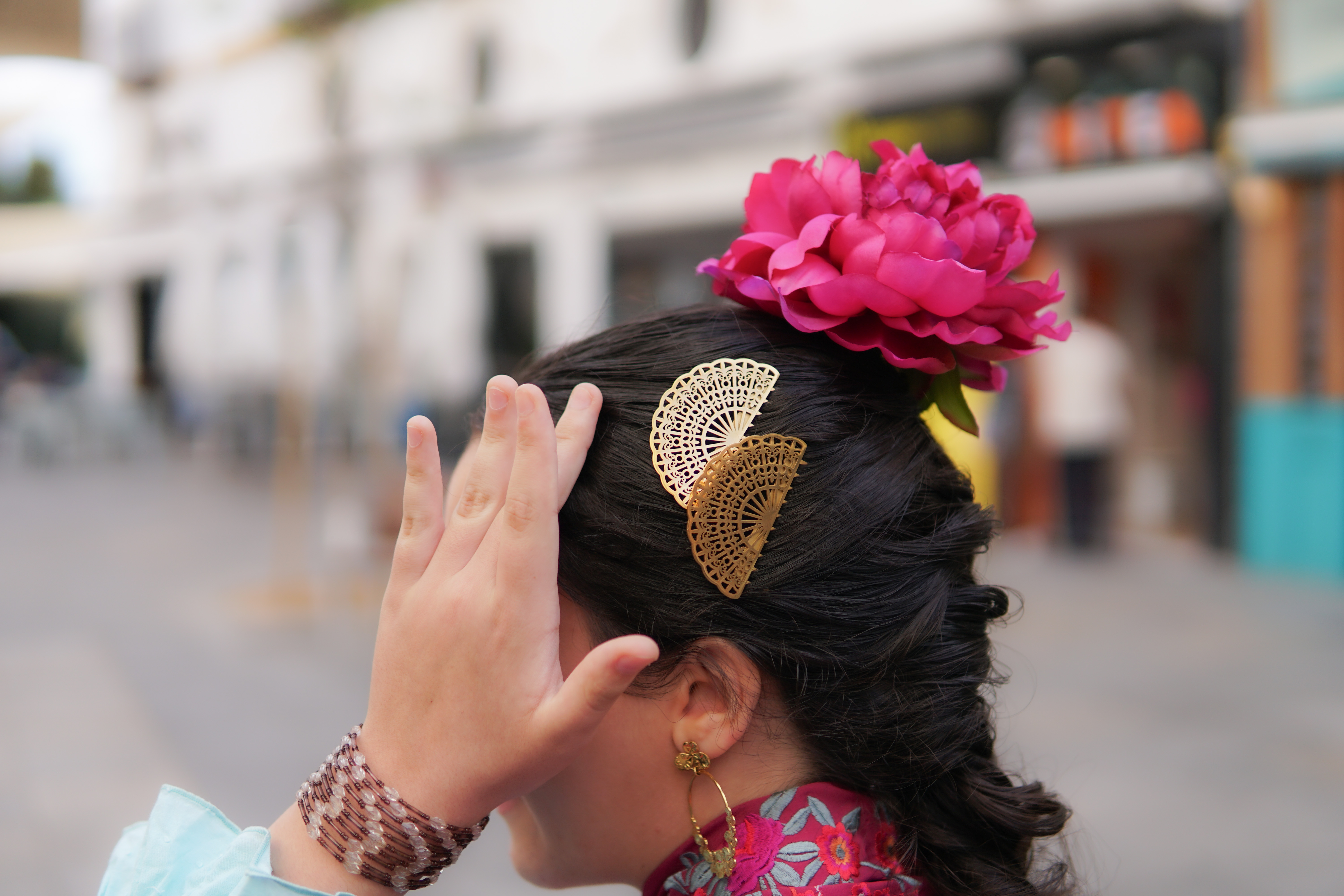 A close-up of the side of a girl's head adorned with traditional hair accessories. She has a large pink flower and two decorative golden fan clips in her dark hair. Her hand is raised, partially obscuring her face, and she is wearing a multi-beaded bracelet on her wrist.