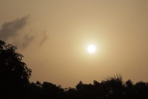 A bright sun is visible in a hazy sky, surrounded by soft clouds.
