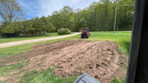 A green lawn with a large open dirt patch that has been flattened. On the far side is a red tractor. A driveway lines the lawn on the left.