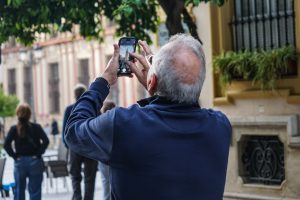 An older man with grey hair is seen from behind, taking a photo of the Giralda tower with his smartphone. He is wearing a dark blue sweater and stands in an outdoor setting with trees and building facades in the background.