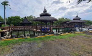 A scenic view of a waterfront area featuring several traditional-style pavilions with thatched roofs, connected by wooden walkways.