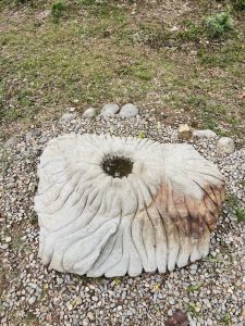 A large, sculpted stone resembling a flower shape is situated on a bed of small pebbles.The stone features a central hollow filled with water, surrounded by wavy, textured edges that give it a natural, artistic appearance. 