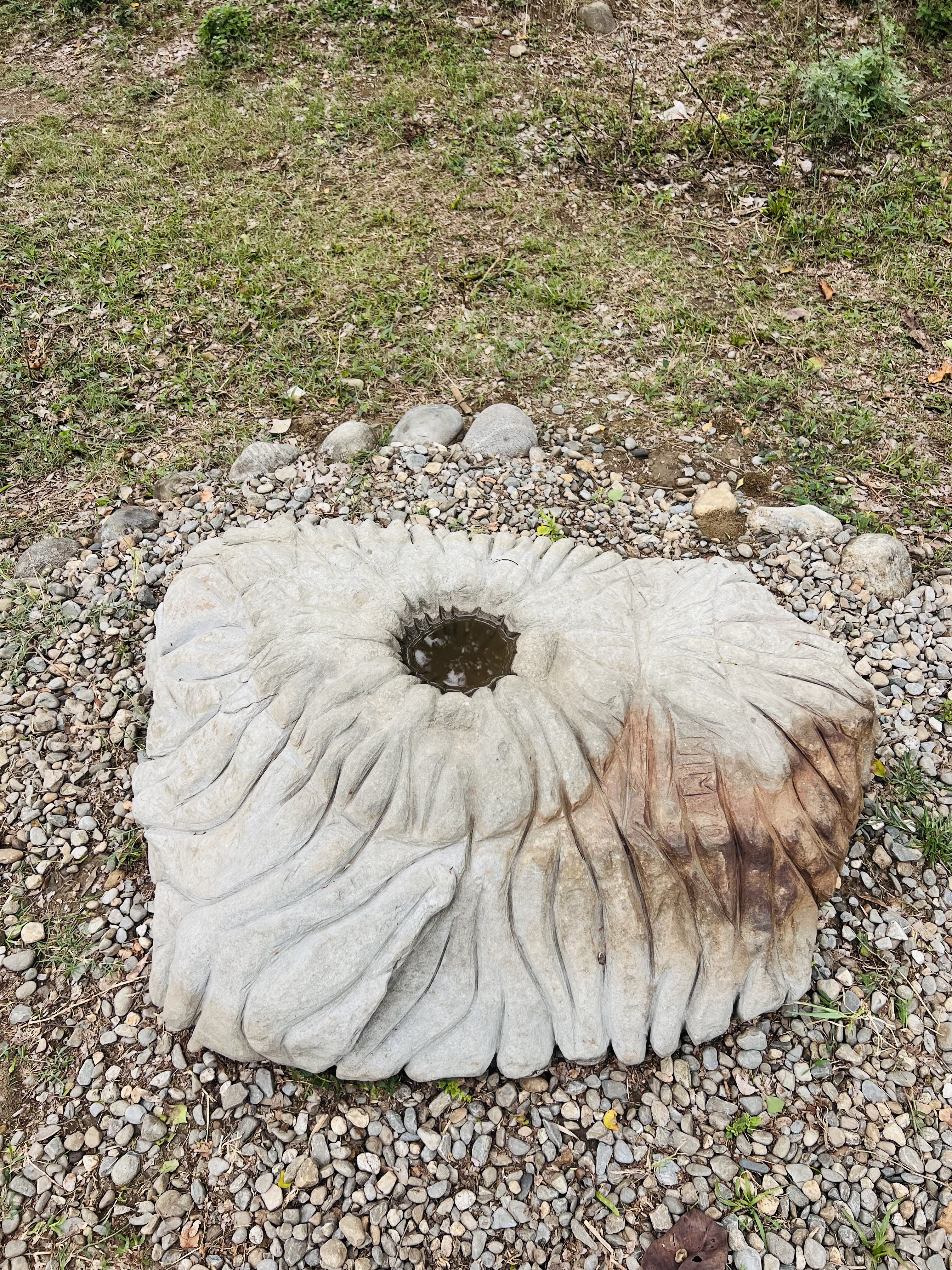 A large, sculpted stone resembling a flower shape is situated on a bed of small pebbles.The stone features a central hollow filled with water, surrounded by wavy, textured edges that give it a natural, artistic appearance.