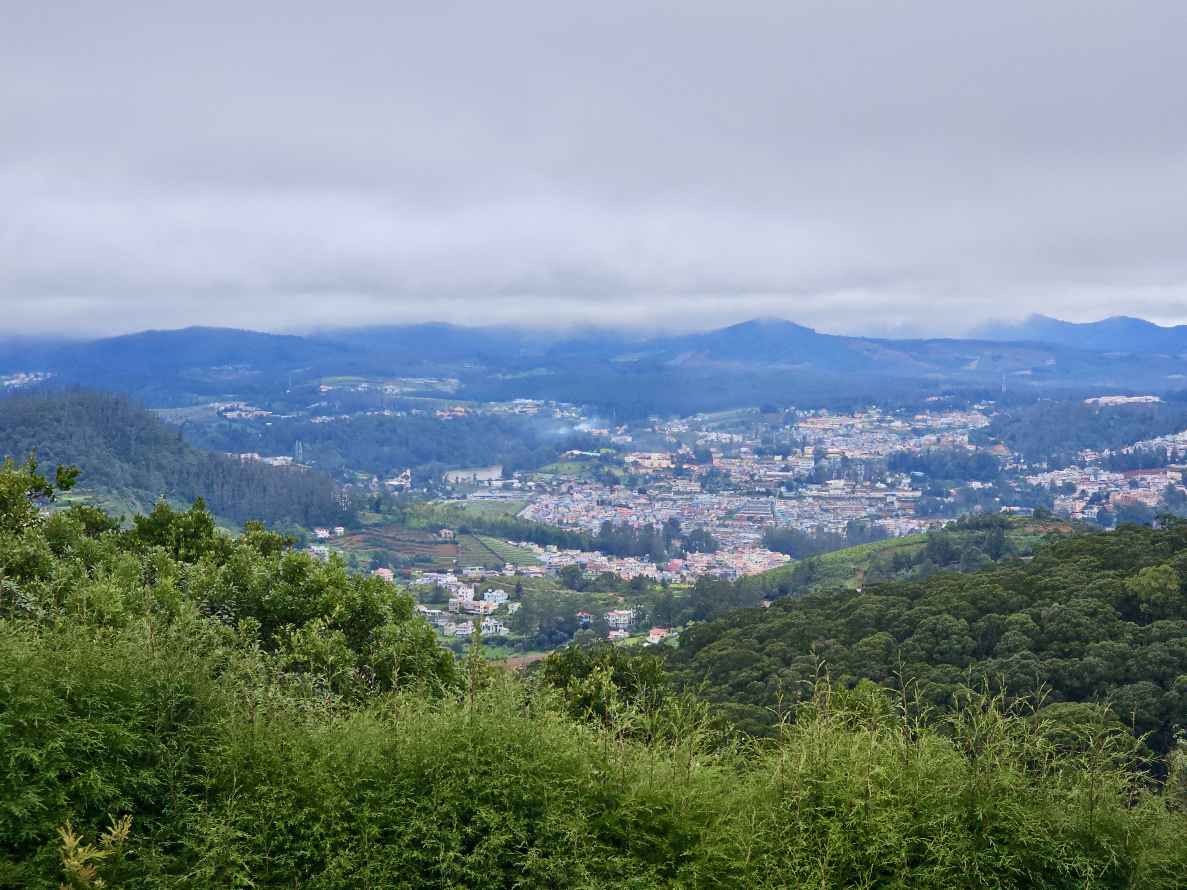 A landscape view of Ooty, a town in the Nilgiri Mountains of Tamil Nadu, India, as seen from Doddabetta peak on a misty and rainy day. The town is nestled among green hills and valleys, with buildings clustered together, and the surrounding landscape is a mix of forests and cultivated land.
