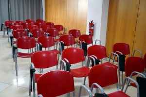 A row of red chairs with metal frames are arranged in a seminar room. The floor is tiled, and a fire extinguisher is mounted on the wall nearby.