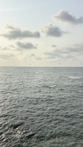 A wide view of the ocean with gentle waves under a partly cloudy sky. Several small boats and a few colorful parasails are visible in the distance, dotting the horizon. The scene captures a peaceful and expansive coastal vista with a soft, muted light.
