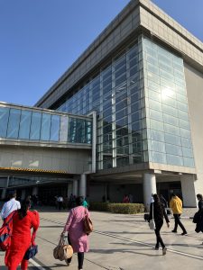 The photo shows people walking toward a modern glass building, likely an airport terminal. The structure has large reflective windows and a skybridge above the entrance. The weather is clear with a bright blue sky, and the sun is shining, casting long shadows on the ground.

