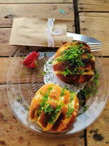 A glass plate with two golden-brown bread rolls topped with parsley sits on a wooden table. A red rose lies beside it. A brown paper bag tied with lace holds cutlery above the plate, blending rustic charm with elegant touches.