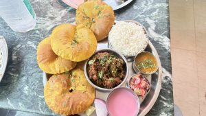 A stainless steel plate containing a meal with several golden-brown puris, a serving of white rice, and a small bowl of a spicy meat dish garnished with cilantro.