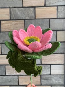 A colorful, handmade crochet flower resembling a pink lotus is held against a backdrop of textured stone bricks.