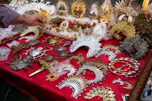 Flea market store showing an assortment of intricate metal ornaments and decorative pieces. There are various styles of crowns and brooches in gold, silver, and bronze, showcasing detailed designs including religious motifs, sun rays, and star patterns