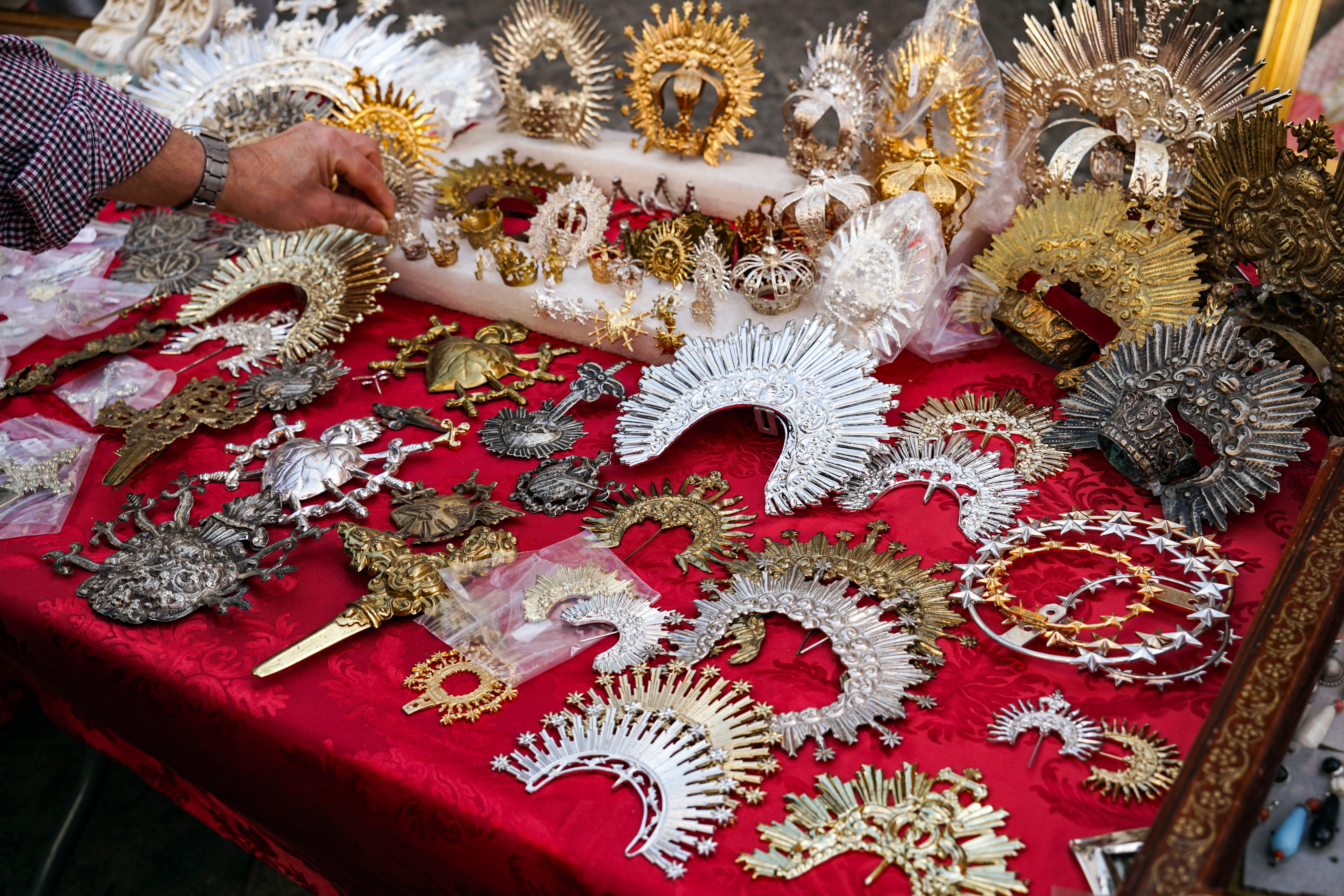 Flea market store showing an assortment of intricate metal ornaments and decorative pieces. There are various styles of crowns and brooches in gold, silver, and bronze, showcasing detailed designs including religious motifs, sun rays, and star patterns