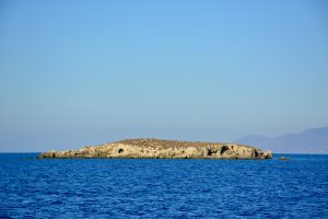 A small, rocky island rising from the calm, blue waters of the sea under a clear, bright sky. Captured from Paphos, Cyprus. 
