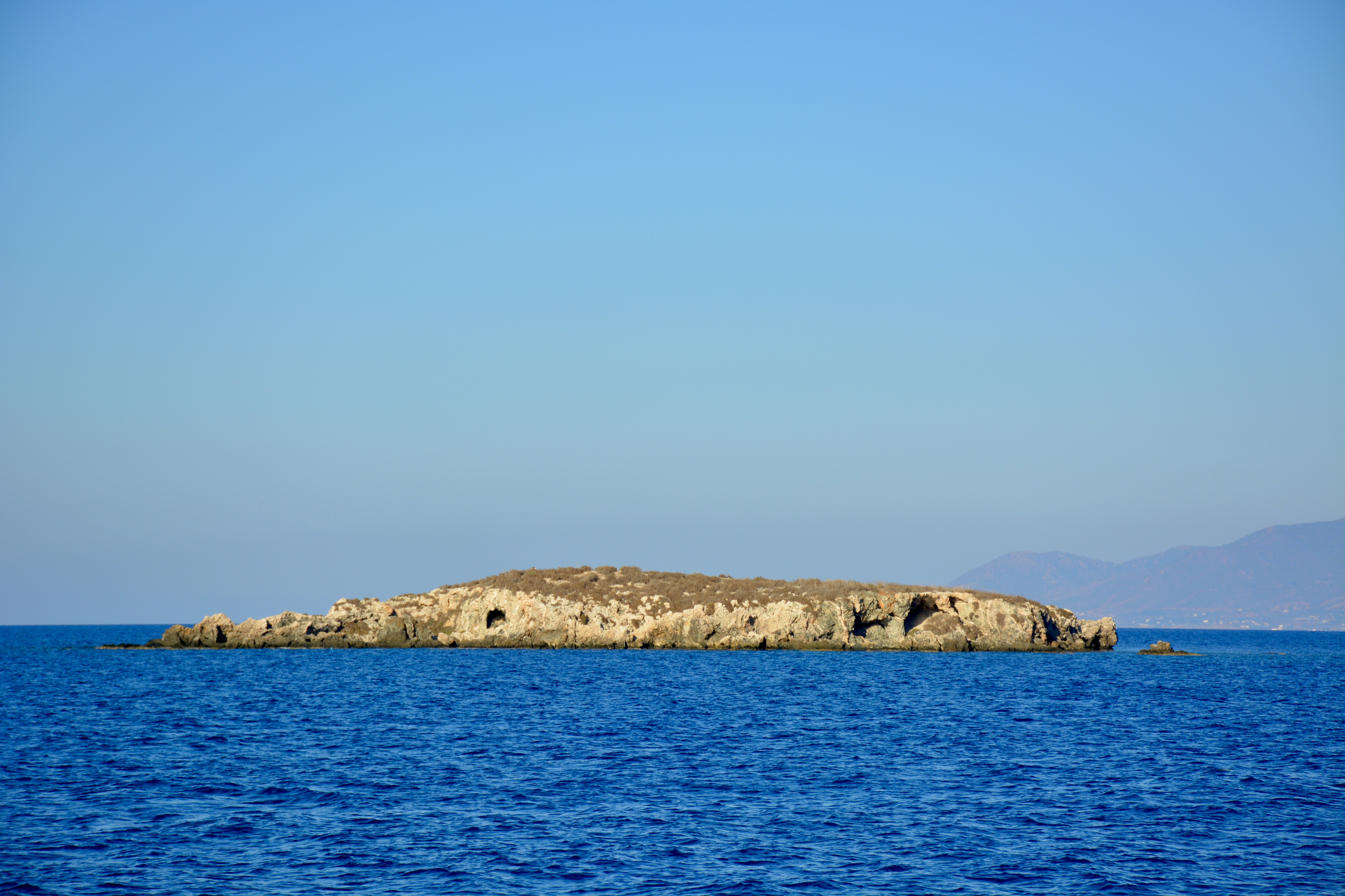 A small, rocky island rising from the calm, blue waters of the sea under a clear, bright sky. Captured from Paphos, Cyprus.