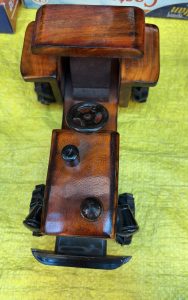 A handcrafted wooden toy tractor with a polished brown finish, featuring a black steering wheel and wheels. The toy is displayed on a bright yellow textured surface.