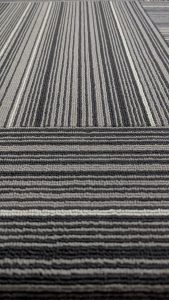 A close-up view of a patterned carpet featuring alternating stripes of black, gray, and white colors. The texture is visible, showcasing a woven appearance with varying thicknesses of the stripes.