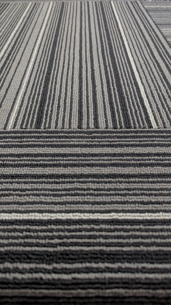 A close-up view of a patterned carpet featuring alternating stripes of black, gray, and white colors. The texture is visible, showcasing a woven appearance with varying thicknesses of the stripes.