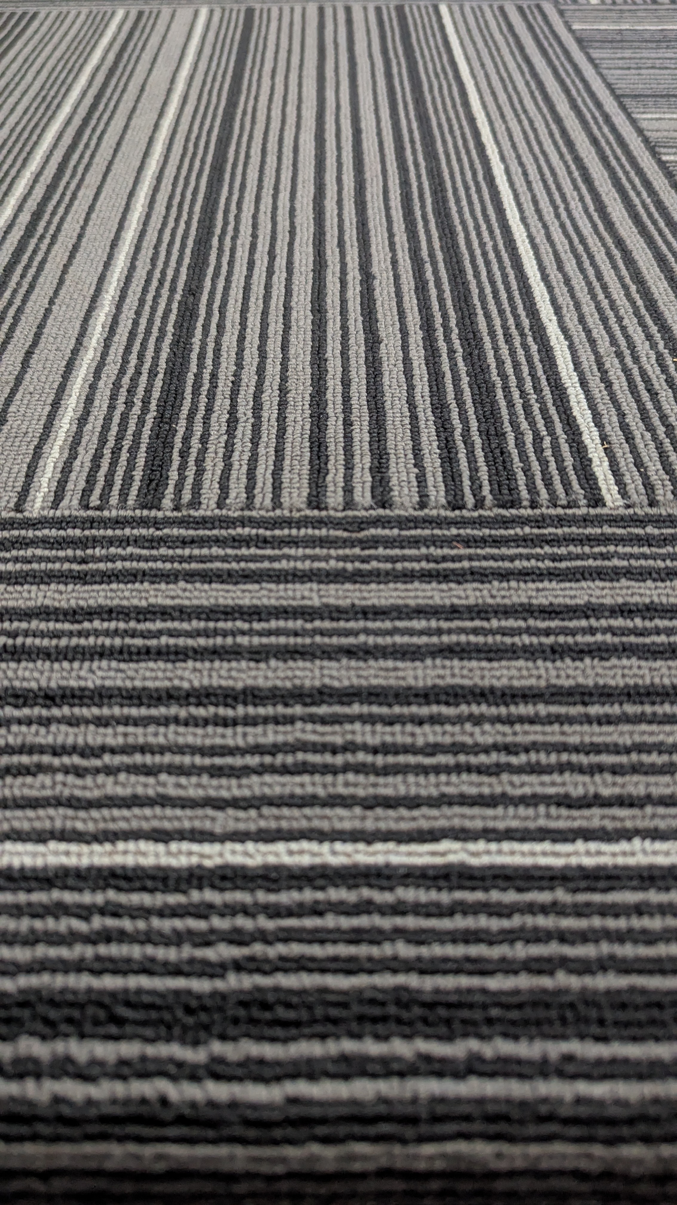 A close-up view of a patterned carpet featuring alternating stripes of black, gray, and white colors. The texture is visible, showcasing a woven appearance with varying thicknesses of the stripes.