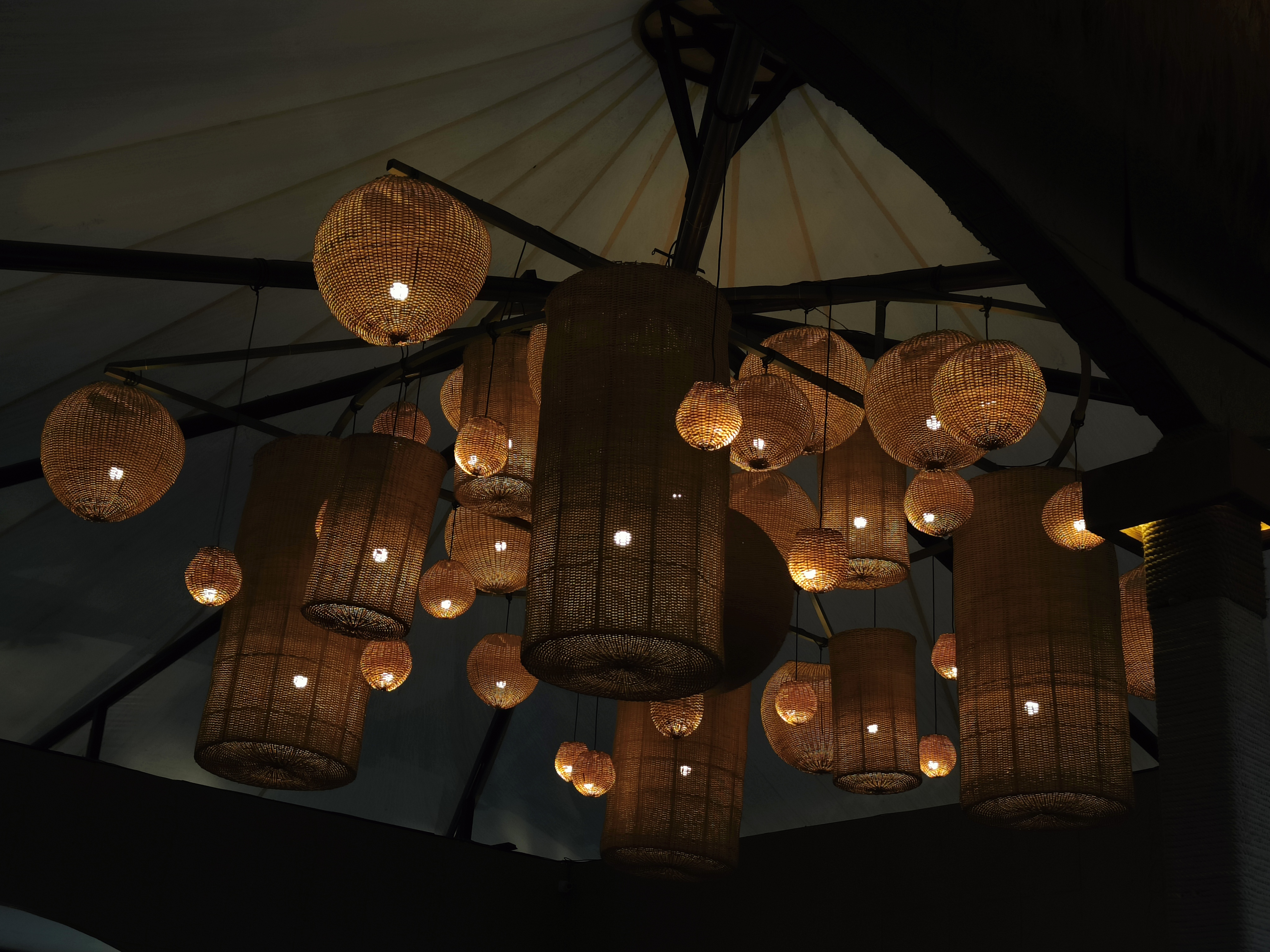 A ceiling adorned with various woven pendant lights, showcasing a mix of round and cylindrical shapes. The lights emit a warm glow against a dark background, with intricate patterns visible in the woven material. 