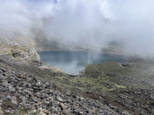 A scenic landscape featuring a tranquil lake surrounded by rocky terrain and grassy patches, partially obscured by fog and clouds.