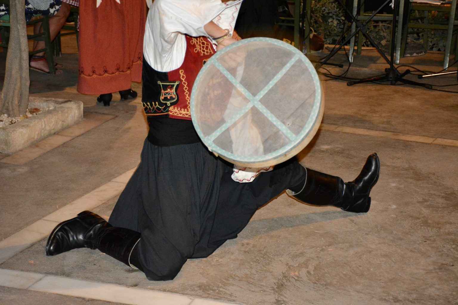 A person in traditional attire kneels with a translucent frame drum in Paphos, Cyprus, during a cultural performance. Another performer is partially visible in the background.