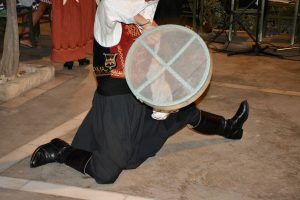A person in traditional attire kneels with a translucent frame drum in Paphos, Cyprus, during a cultural performance. Another performer is partially visible in the background.