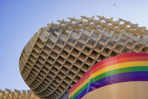 Modern architectural structure (Metropol Parasol) featuring a curved and layered design with a series of protruding elements. The structure is partially covered by a rainbow flag, emphasizing colors of the LGBTQ+ pride flag. The background shows a clear blue sky with a few swifts flying.
