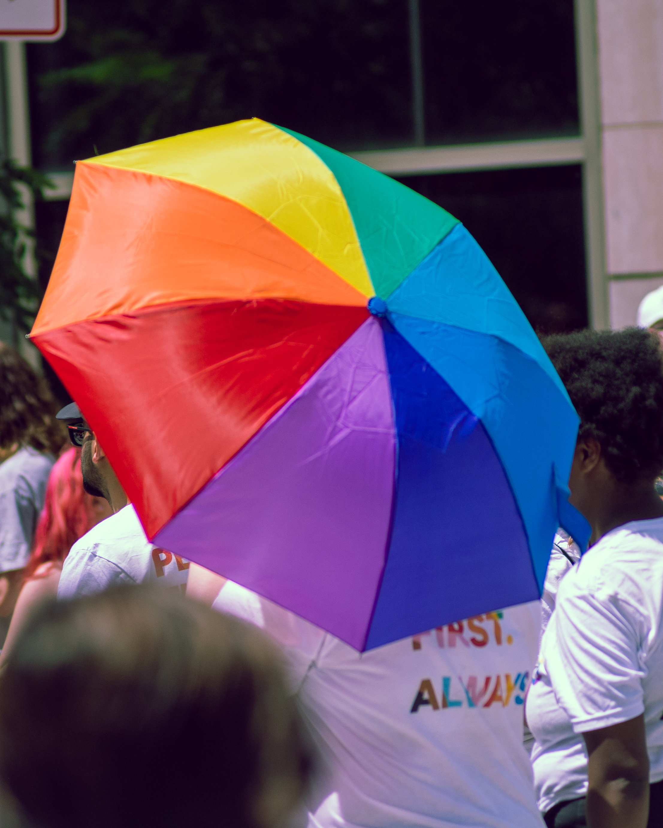 A vibrant rainbow umbrella shades a person in a white t-shirt with colorful text during a Pride event. They are surrounded by others in festive attire, with sunlight casting bright reflections off the umbrella’s surface.