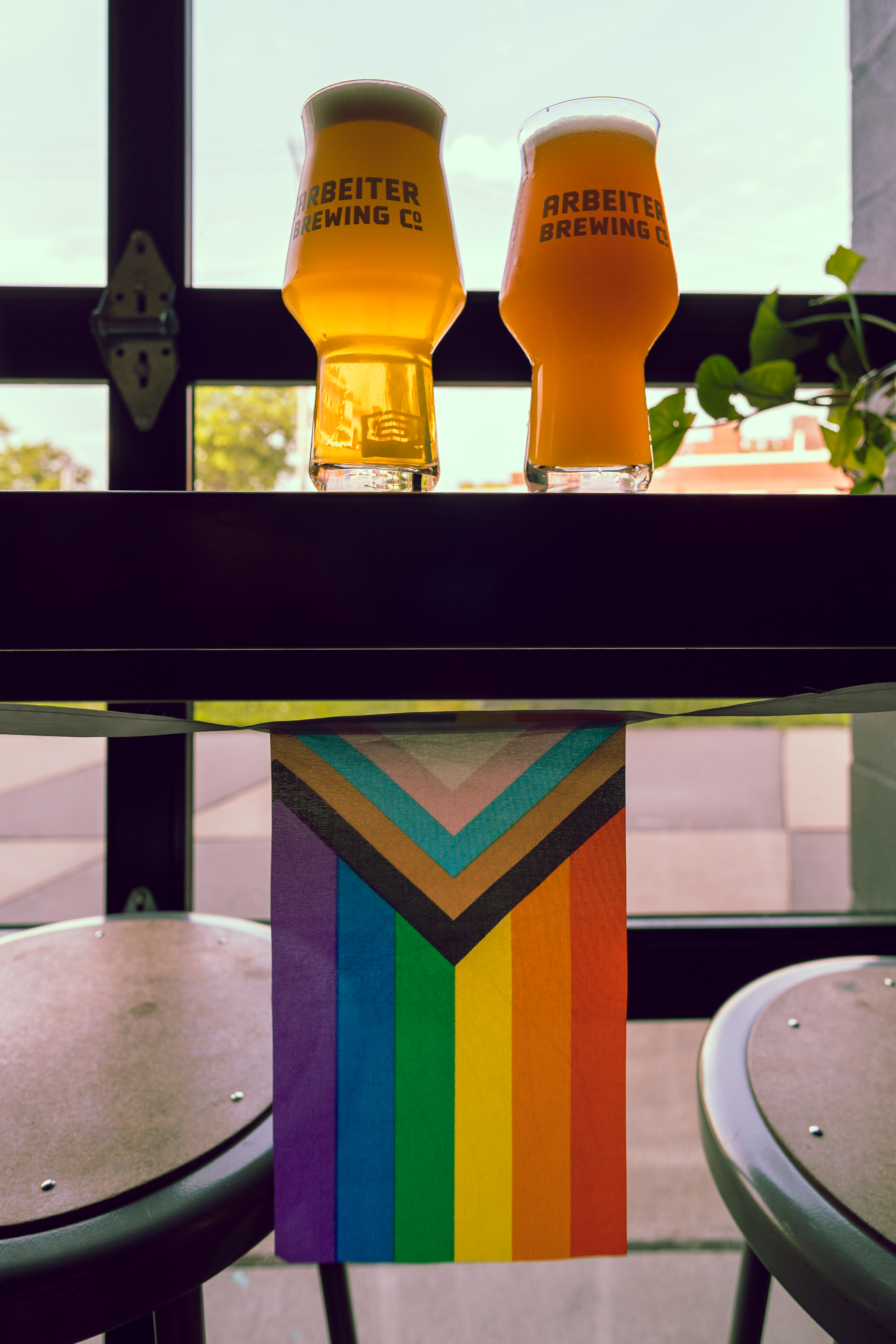 Two beers sit on a ledge in front of a glass garage door with a Progress Pride flag hanging from the bottom of the ledge.
