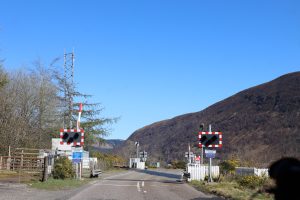 A rural road leading to a railway crossing, surrounded by trees and hills. 
