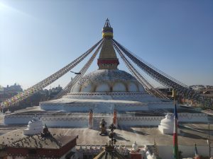 

A large bouddhanath stupa with a white dome and colorful prayer flags draped from the top, set against a clear blue sky.