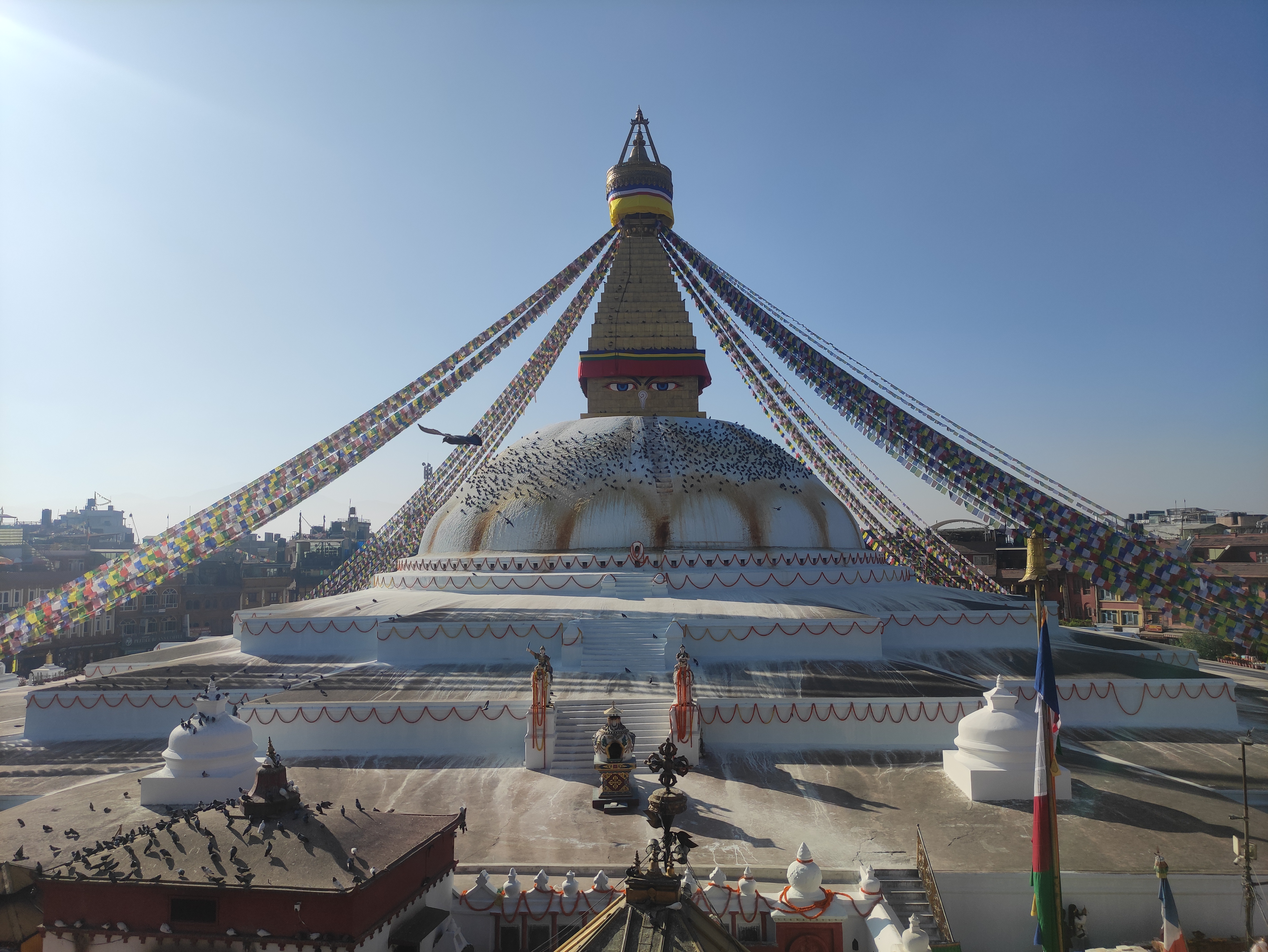 A large bouddhanath stupa with a white dome and colorful prayer flags draped from the top, set against a clear blue sky.