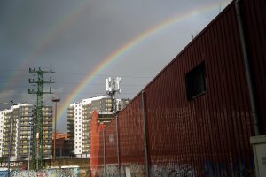 A vibrant double rainbow arcs across a cloudy sky above a cityscape. In the foreground, a red corrugated metal wall is partially visible, adorned with graffiti. Behind it, several modern buildings with a mix of gray and white facades stand, some featuring yellow balconies. 