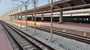 A train station platform with metal supports and a roof is visible, featuring several train carriages on the adjacent tracks. 