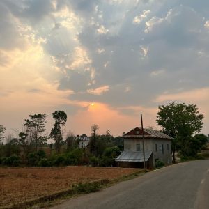 A rural landscape at sunset featuring a winding road on the right side. In the foreground, there is a simple, two-story house with a metal roof, partially obscured by trees