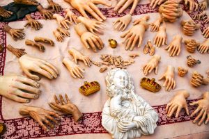 A variety of hand sculptures and figures are arranged on a light fabric surface, with a focus on both large and small hands in different poses and styles. Among them is a white figurine of a mother holding a child, flanked by two small golden crowns. 